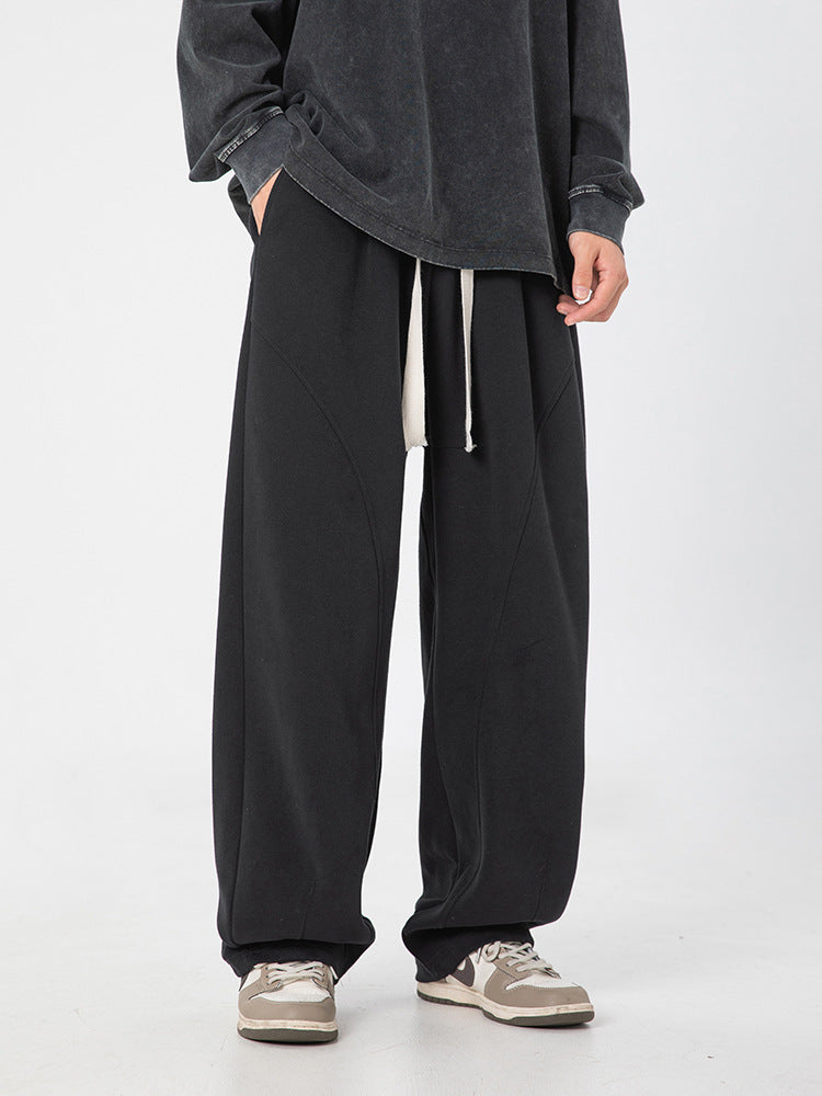 A model is wearing oversized black wide-leg sweatpants with diagonal seams and drawstring waist, paired with a dark sweatshirt and gray sneakers