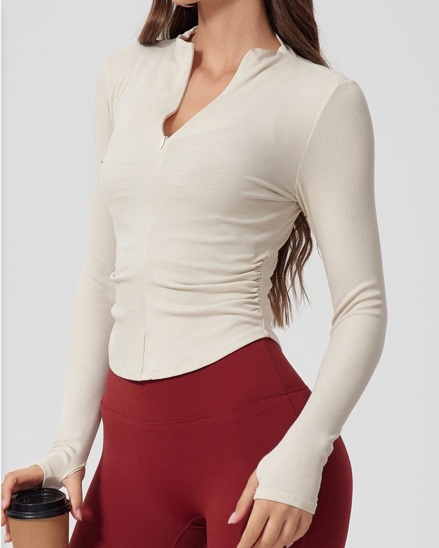 A woman wearing a cream ribbed long-sleeve top with stand collar and v-neck, paired with burgundy leggings, holding coffee cup against light background