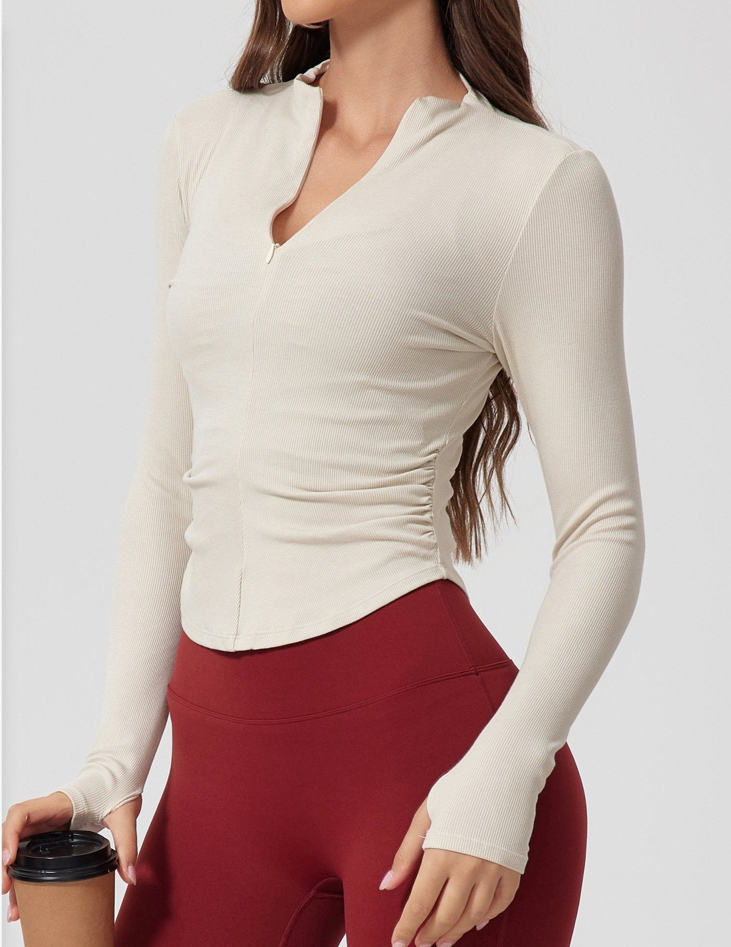 A woman wearing a cream ribbed long-sleeve top with stand collar and v-neck, paired with burgundy leggings, holding coffee cup against light background