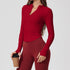 A woman wearing a red ribbed long-sleeve top with v-neck collar and side ruching, paired with burgundy leggings, holding coffee cup against neutral background