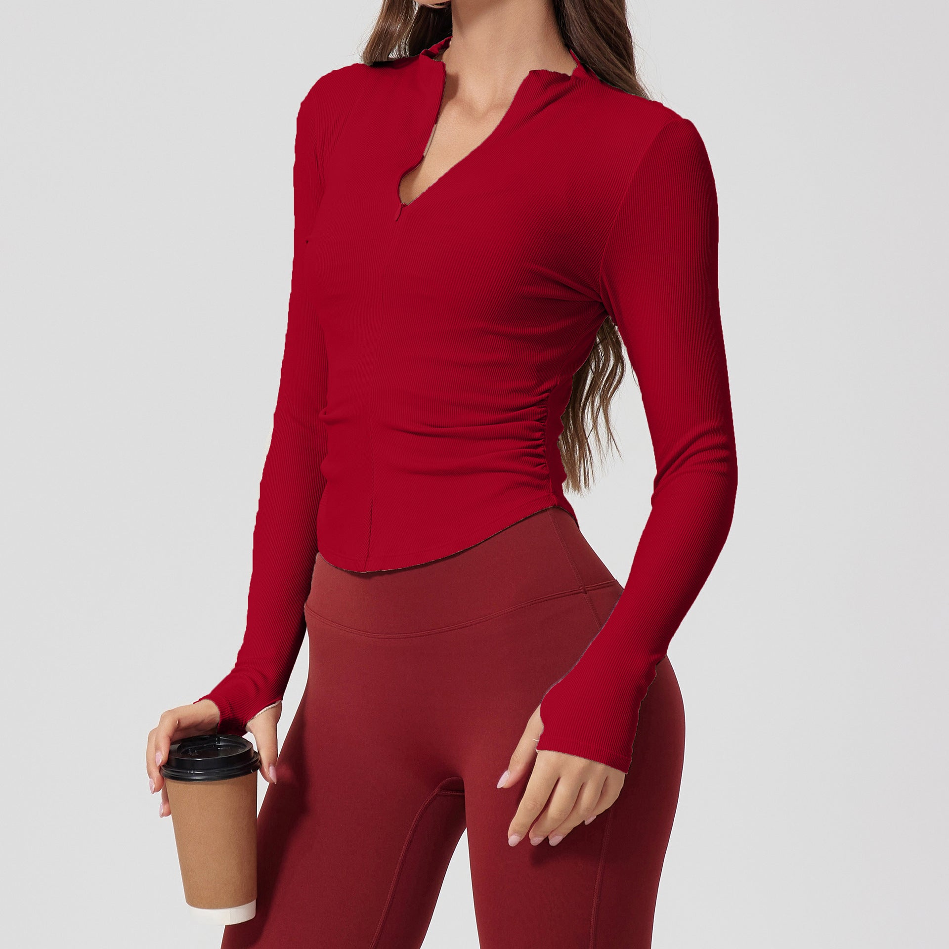 A woman wearing a red ribbed long-sleeve top with v-neck collar and side ruching, paired with burgundy leggings, holding coffee cup against neutral background