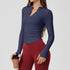 A woman wearing a navy blue ribbed long-sleeve top with v-neck collar and side ruching, paired with burgundy leggings, holding coffee cup against neutral background