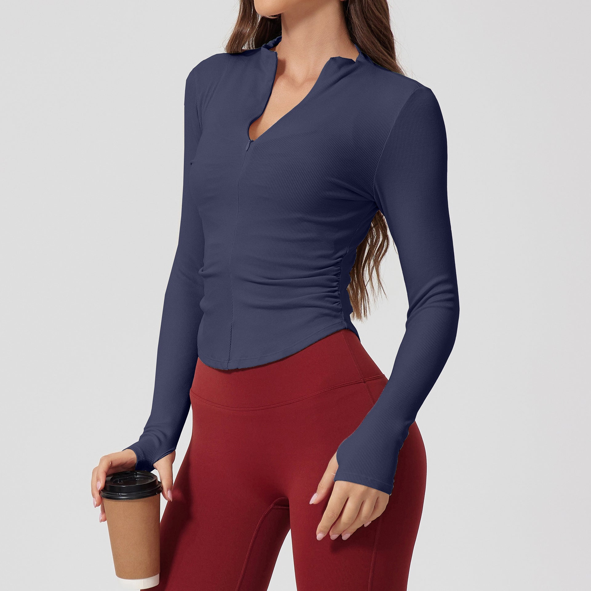 A woman wearing a navy blue ribbed long-sleeve top with v-neck collar and side ruching, paired with burgundy leggings, holding coffee cup against neutral background
