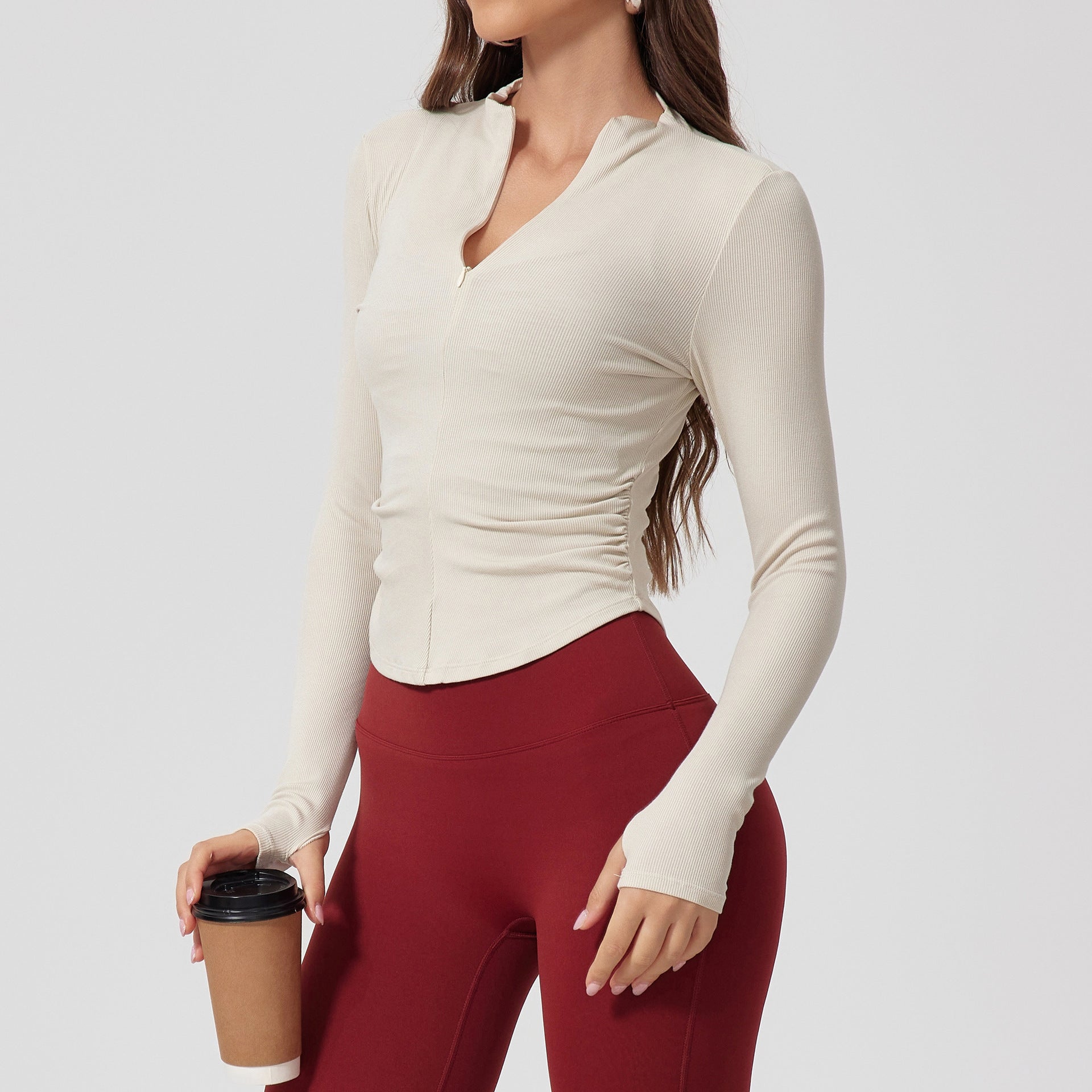 A woman wearing a cream ribbed long-sleeve top with v-neck collar and side ruching, paired with burgundy leggings, holding coffee cup against light background