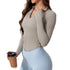 A woman wearing a light beige ribbed long-sleeve top with v-neck collar and side ruching, paired with light blue leggings, holding coffee cup against light background