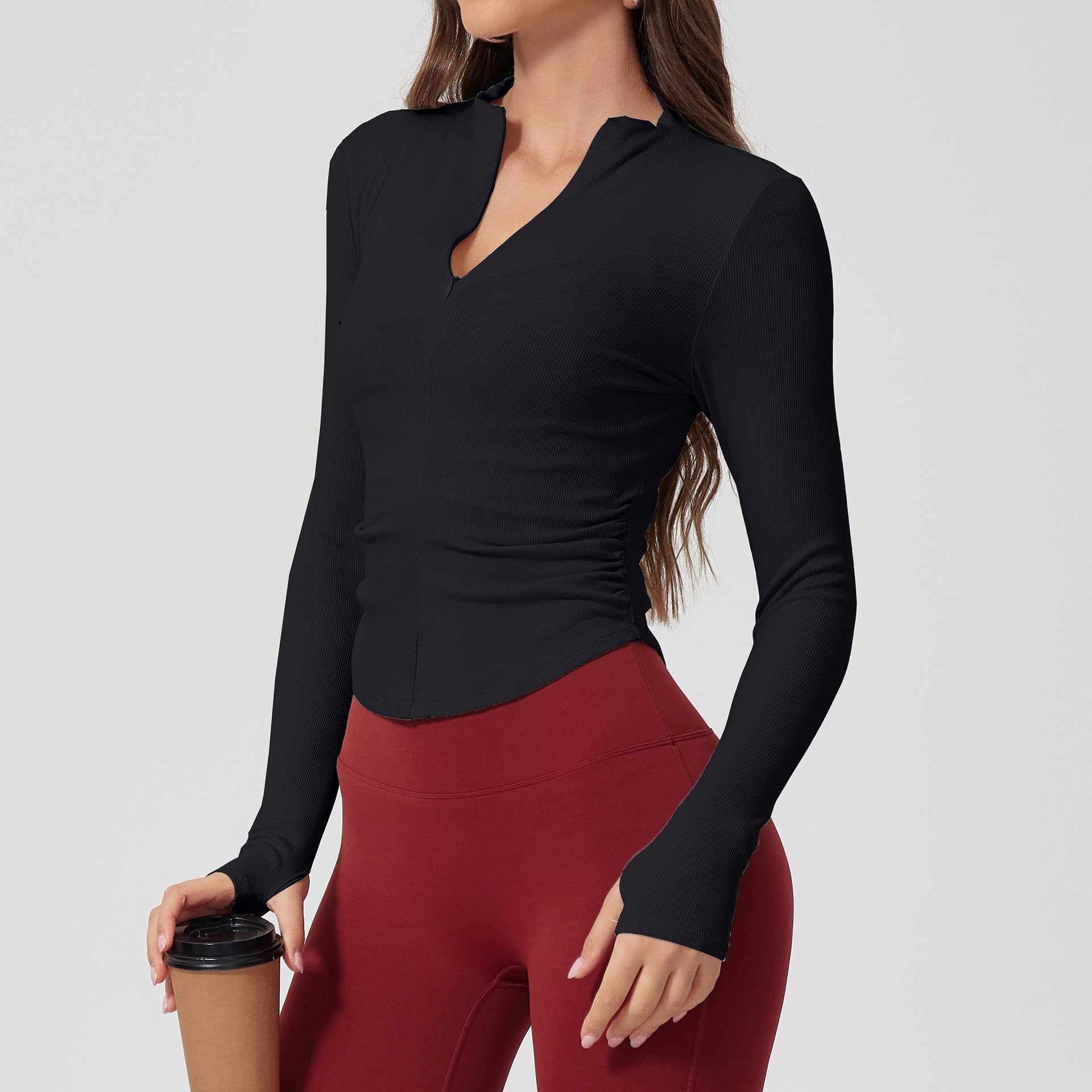 A woman wearing a black ribbed long-sleeve top with v-neck collar and side ruching, paired with burgundy leggings, holding coffee cup against neutral backgroun