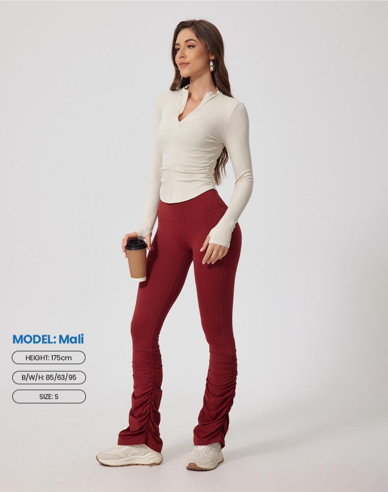 A woman standing in cream ribbed top with v-neck collar and burgundy ruched leggings, holding coffee cup against white background. Model Mali, 175cm, size S