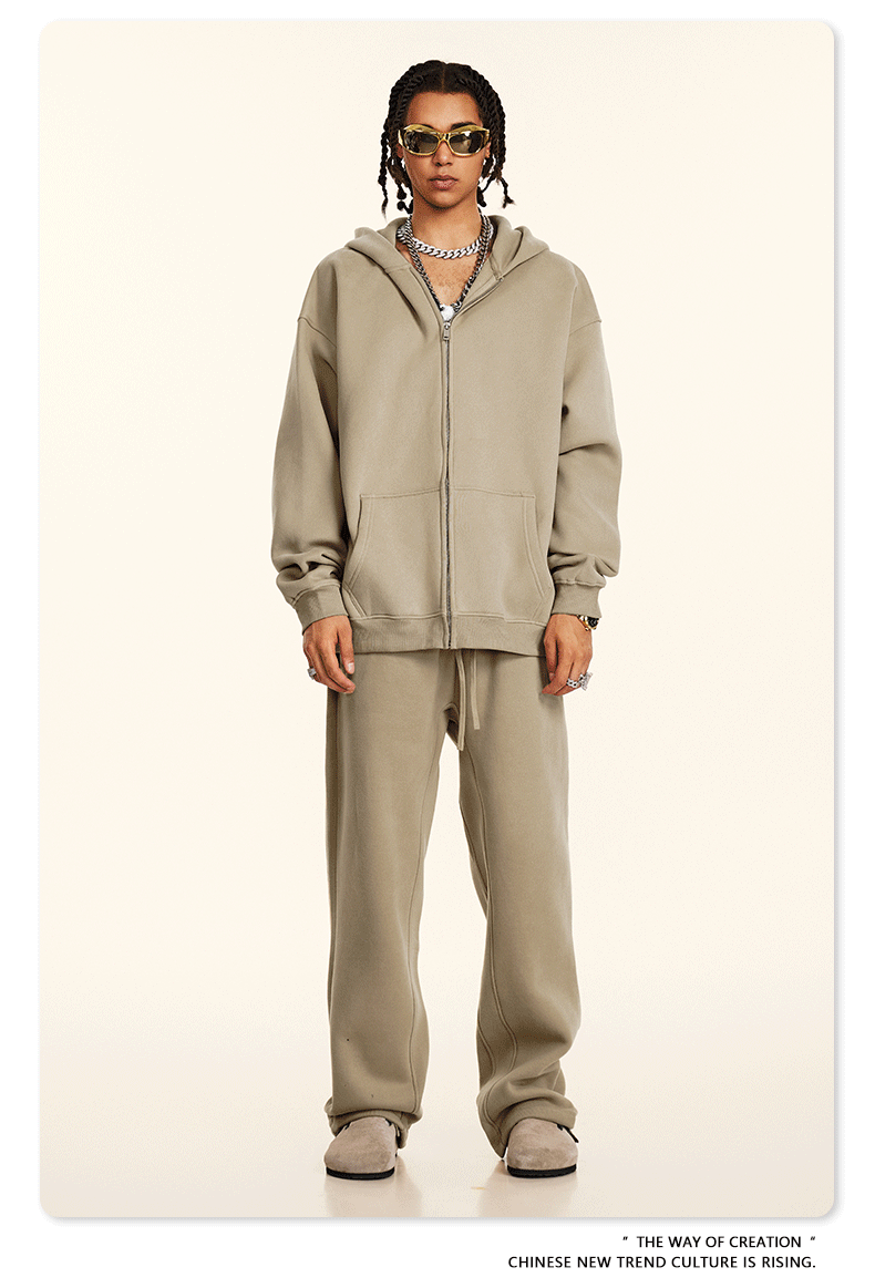 Model wearing beige oversized zip-up hoodie and matching sweatpants with gold-framed sunglasses and layered silver chain necklaces against cream background