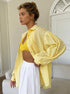 A woman wearing a yellow eyelet oversized shirt over a yellow crop top paired with white linen pants standing against a light couch backdrop