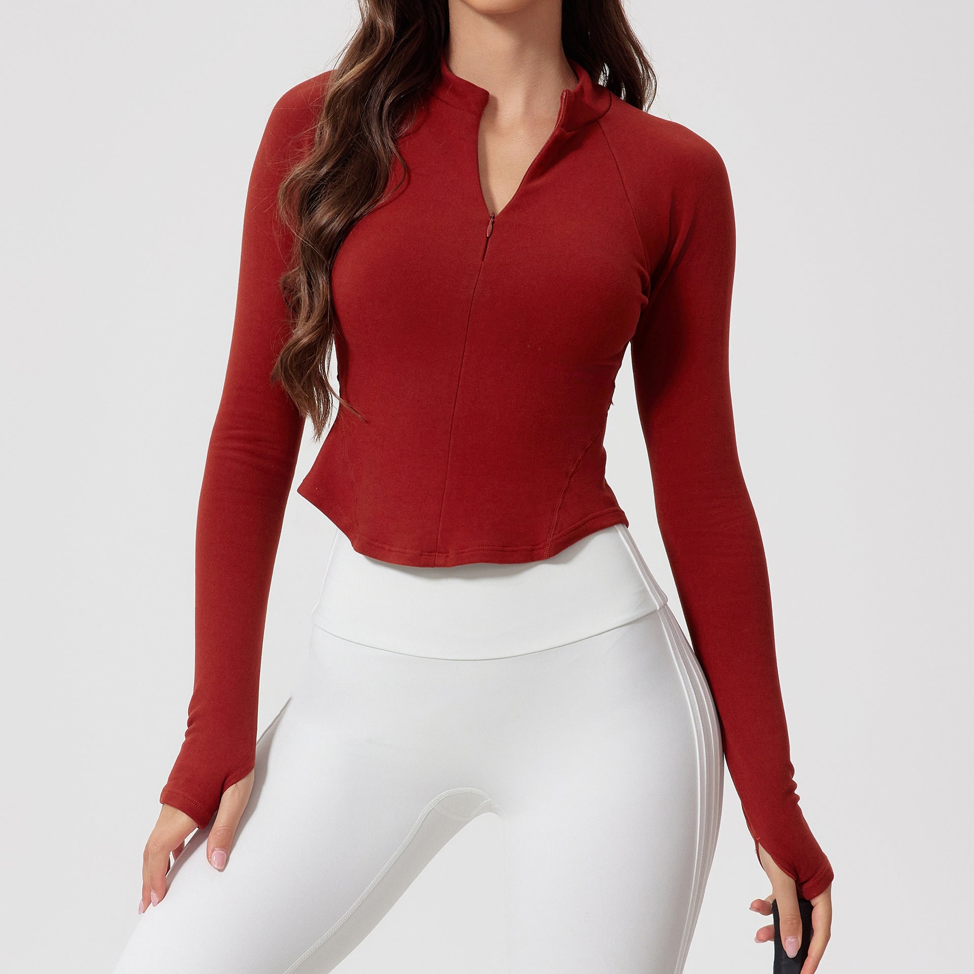 A model is wearing a rust red mandarin collar crop top with white leggings. Long sleeves feature thumb holes for active wear functionality