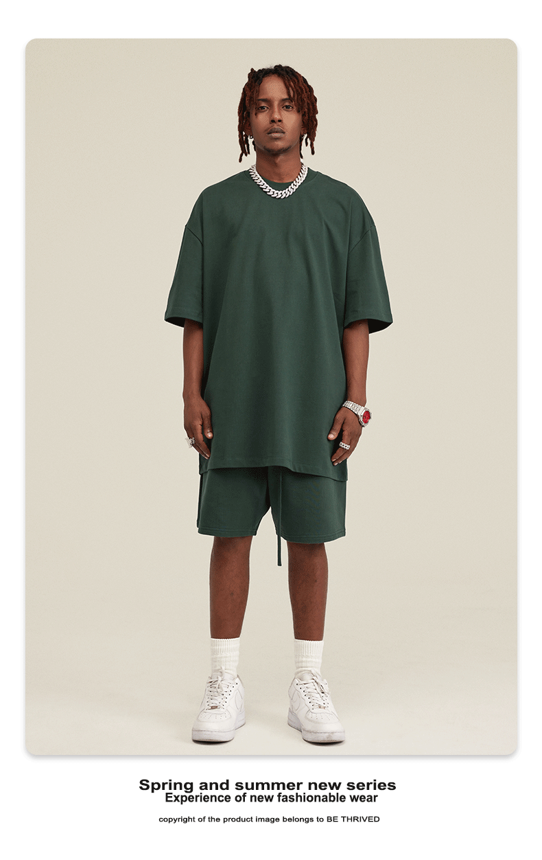 Model with dreadlocks wearing forest green oversized t-shirt and matching shorts with silver jewelry and white sneakers against cream background