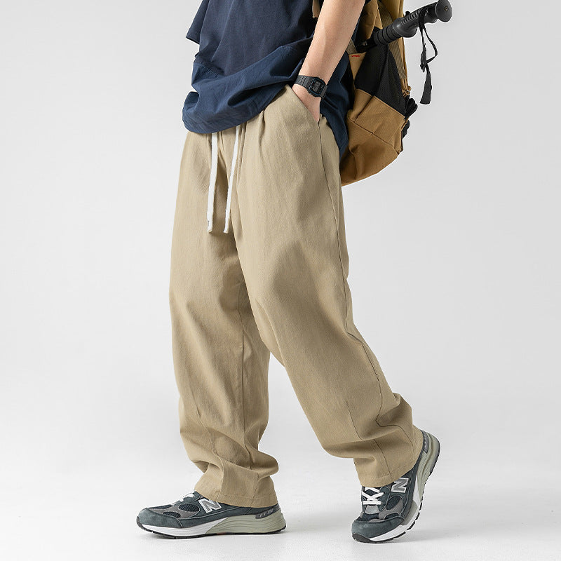 A model is wearing beige wide-leg drawstring pants, a navy oversized T-shirt, and gray sneakers, standing with one hand in the pocket and carrying a backpack