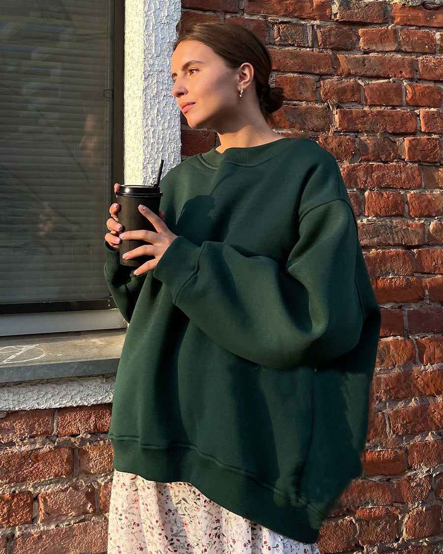 Forest green oversized sweatshirt styled with floral print skirt and holding coffee cup against brick wall