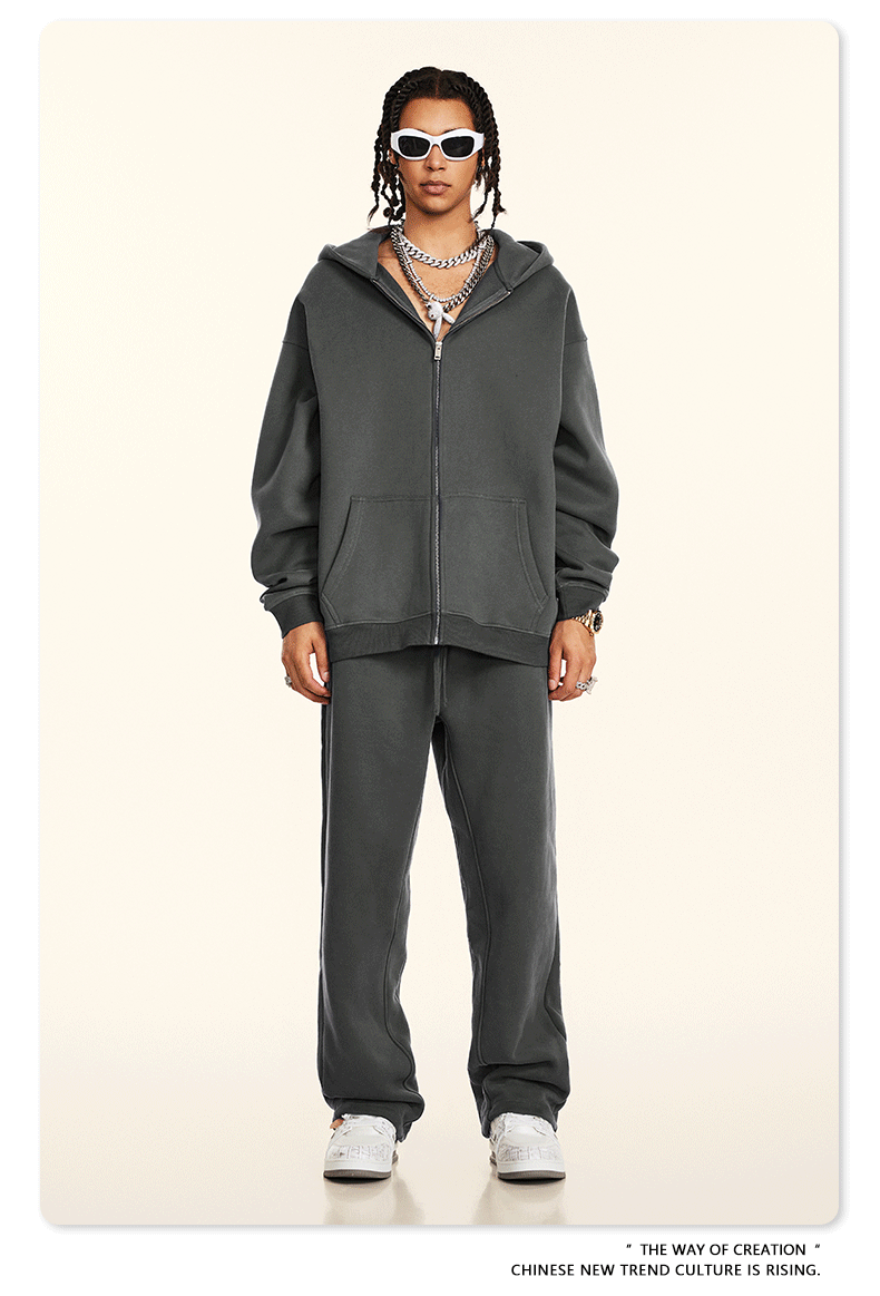 Model wearing dark gray oversized zip-up hoodie and matching sweatpants with white rectangular sunglasses and silver chain accessories against cream backdrop