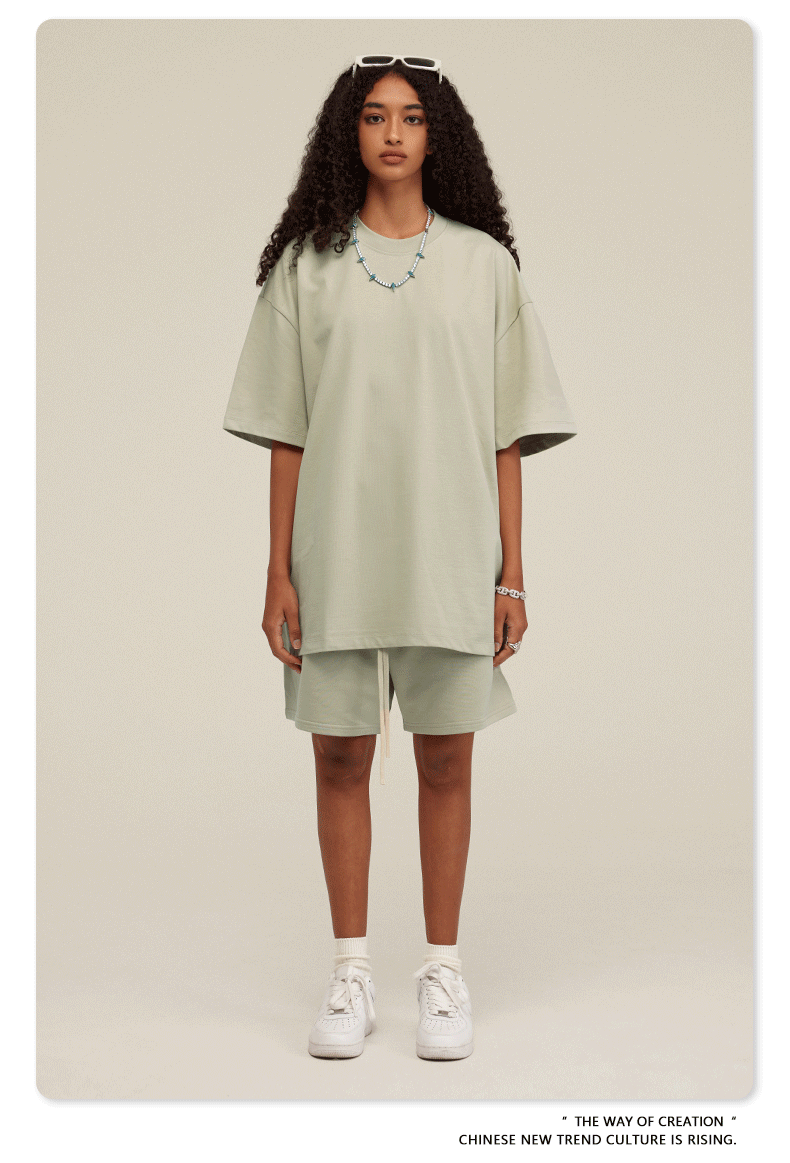 A woman with curly hair wearing a light mint green oversized t-shirt and matching shorts set, styled with white socks, sneakers, and a bead necklace