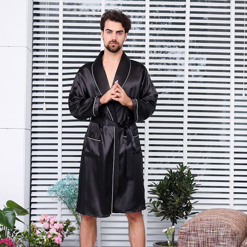 Man in black silk robe with white piping detail standing with hands clasped, against white blinds backdrop with decorative indoor plants and contemporary home styling