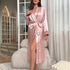 A woman in a long pink silk robe with black trim and tie waist, standing in a sunlit room