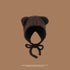 A chocolate brown teddy ear hat with chin ties against brown background, featuring rounded ears and soft plush exterior with textured finish