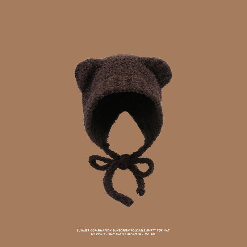 A chocolate brown teddy ear hat with chin ties against brown background, featuring rounded ears and soft plush exterior with textured finish