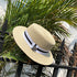 A beige straw boater hat resting on a black metal fence, surrounded by green palm leaves, showcasing the ribbon and charm detail.