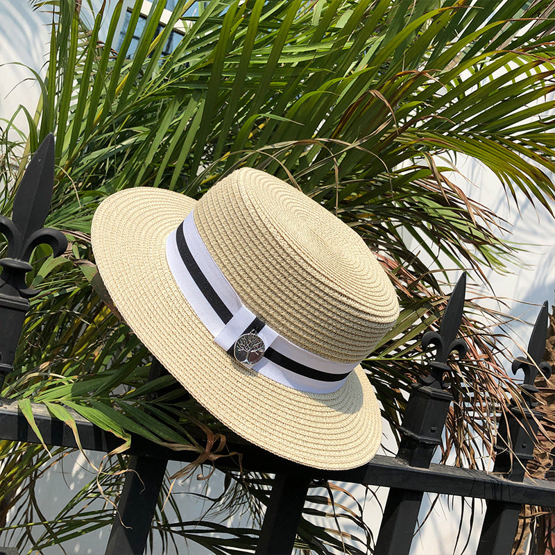 A beige straw boater hat resting on a black metal fence, surrounded by green palm leaves, showcasing the ribbon and charm detail.