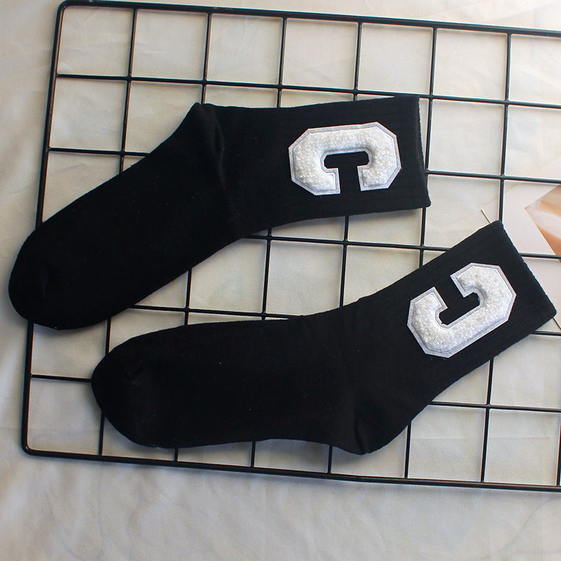 A pair of black varsity-style crew socks with white textured "C" patches is laid out on a black wire grid over a neutral fabric surface