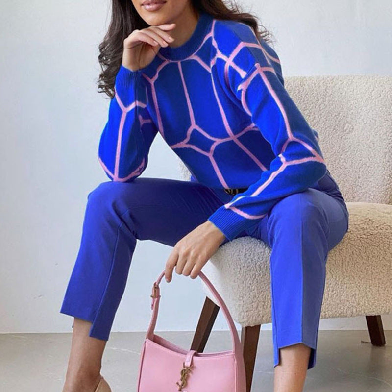 A model is sitting on beige chair wearing royal blue geometric sweater with pink pattern, matching pants, and holding a pink handbag