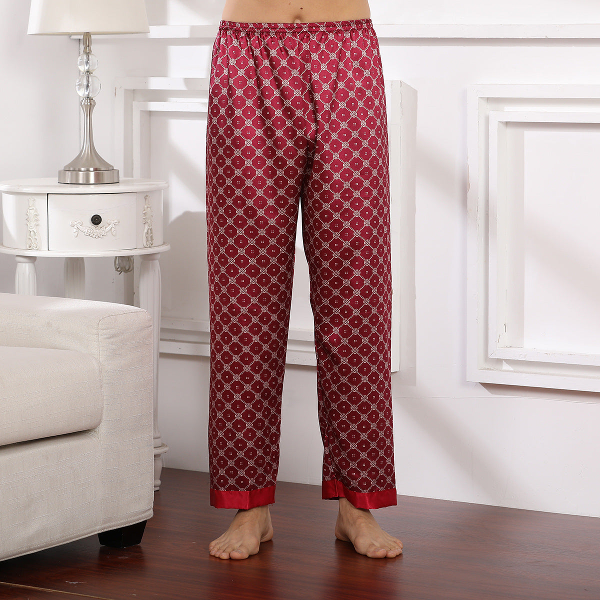 Model wearing deep red silk pajama pants with repeating geometric pattern and red trim at bottom, standing barefoot on wooden floor
