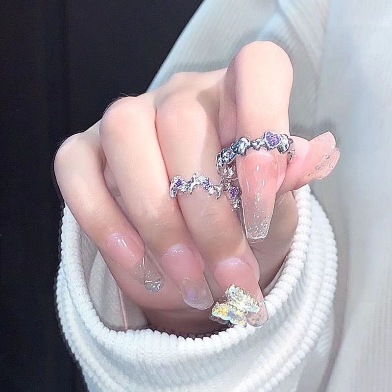 A person with clear manicured nails holding white fabric, wearing silver rings with purple accents and star design, showcasing sparkly nail art