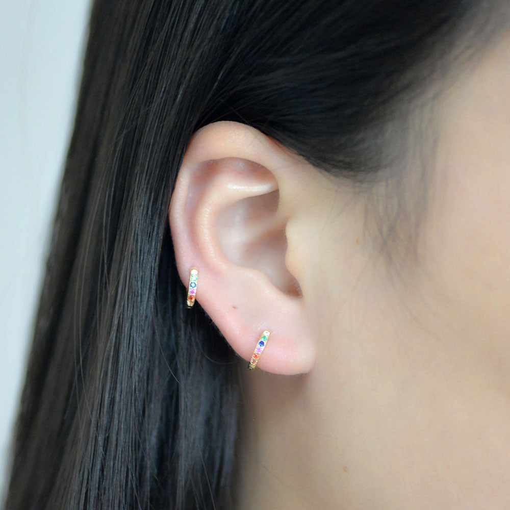 A woman wearing two gold rainbow huggie hoop earrings in her ear, close-up against black hair