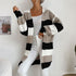 A woman wearing black and taupe colorblock cardigan with white cami top, styled with white pants and layered chain necklaces in home setting