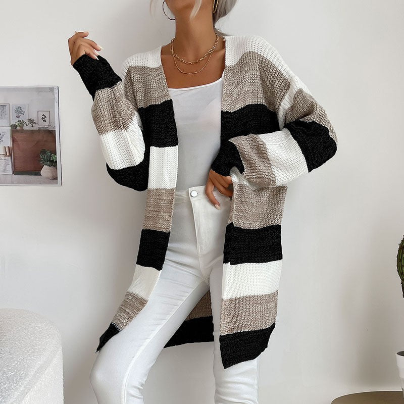 A woman wearing black and taupe colorblock cardigan with white cami top, styled with white pants and layered chain necklaces in home setting