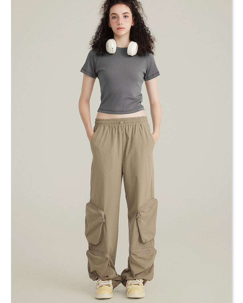Model with curly dark hair wearing a gray crop top, beige cargo pants with zippered pockets, white headphones around neck on neutral background