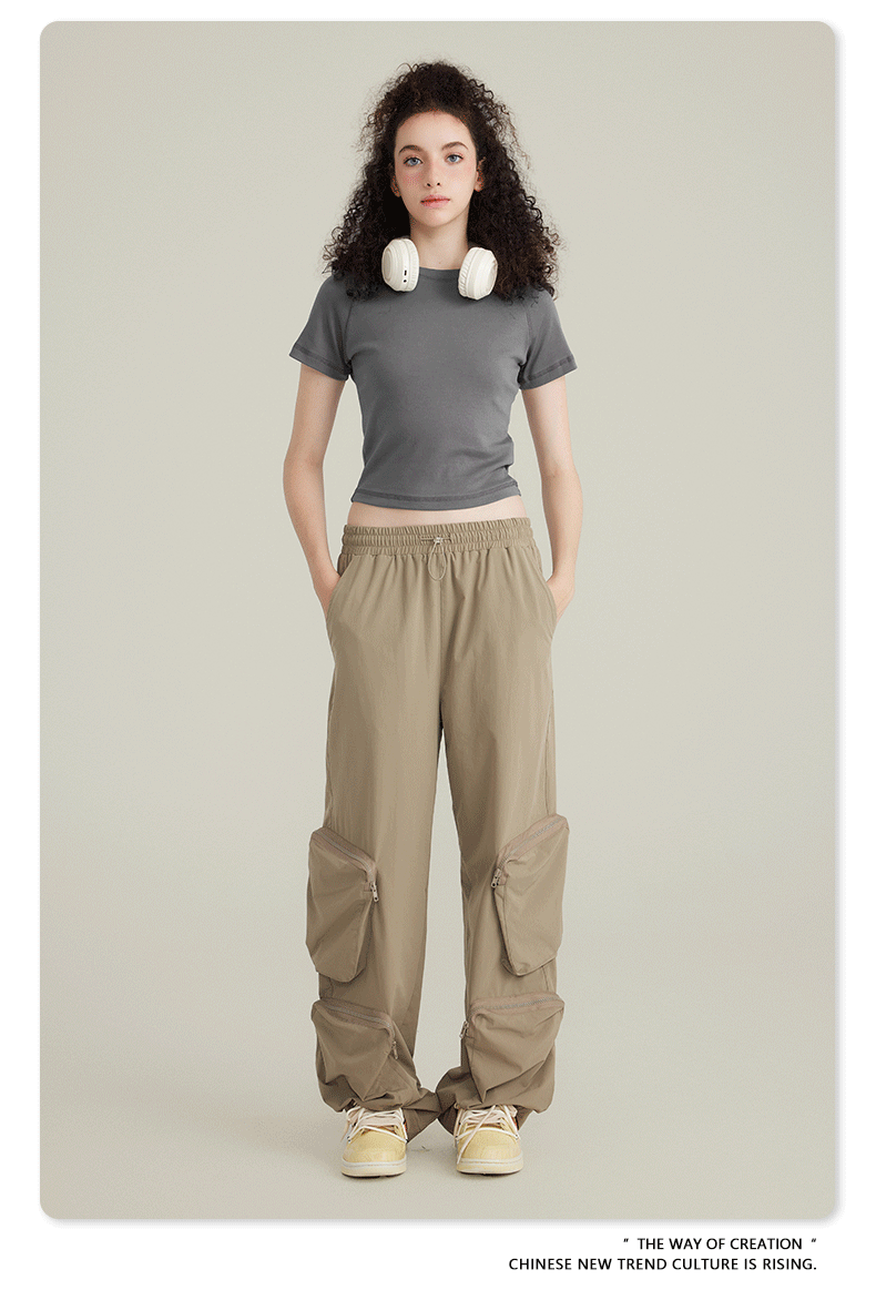 Model with curly dark hair wearing a gray crop top, beige cargo pants with zippered pockets, white headphones around neck on neutral background