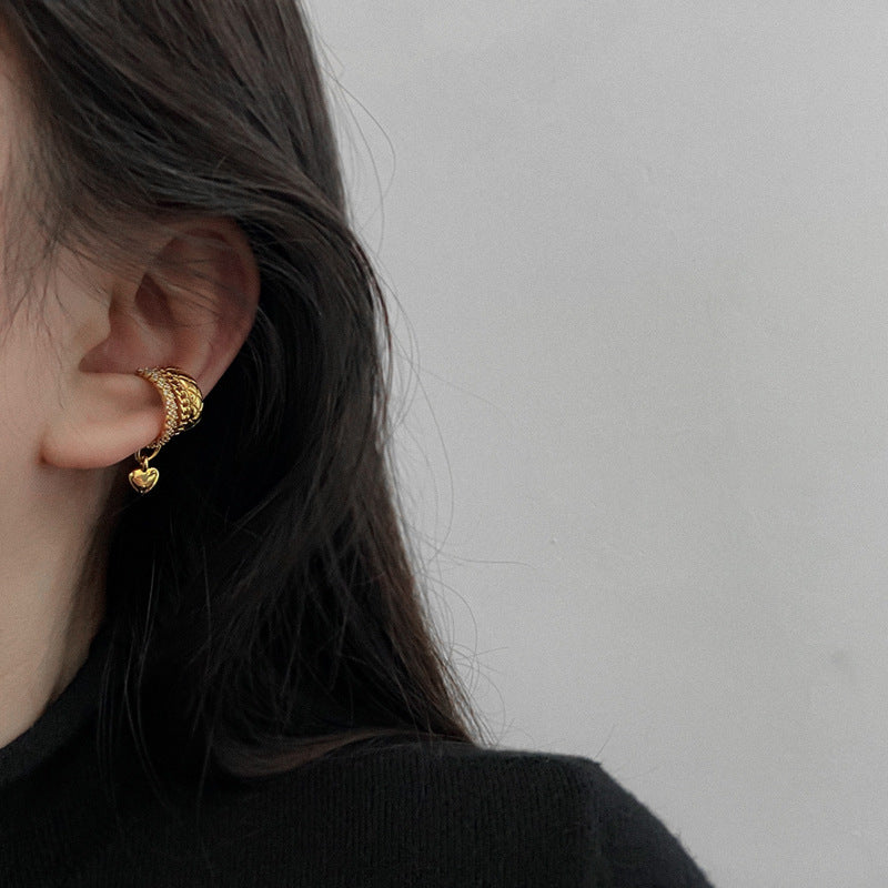 Gold ear cuff with heart charm worn on ear against dark hair and black clothing