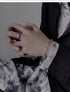 A person with crossed hands wearing a silver chain bracelet with a cross pendant and a silver ring with a blue gemstone, styled with a dark tie-dye patterned shirt.