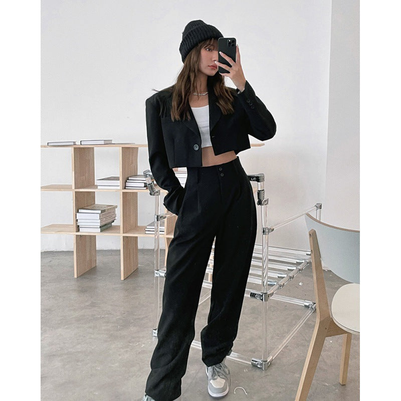 Woman in black cropped blazer and wide-leg pants taking mirror selfie, wearing beanie and white crop top