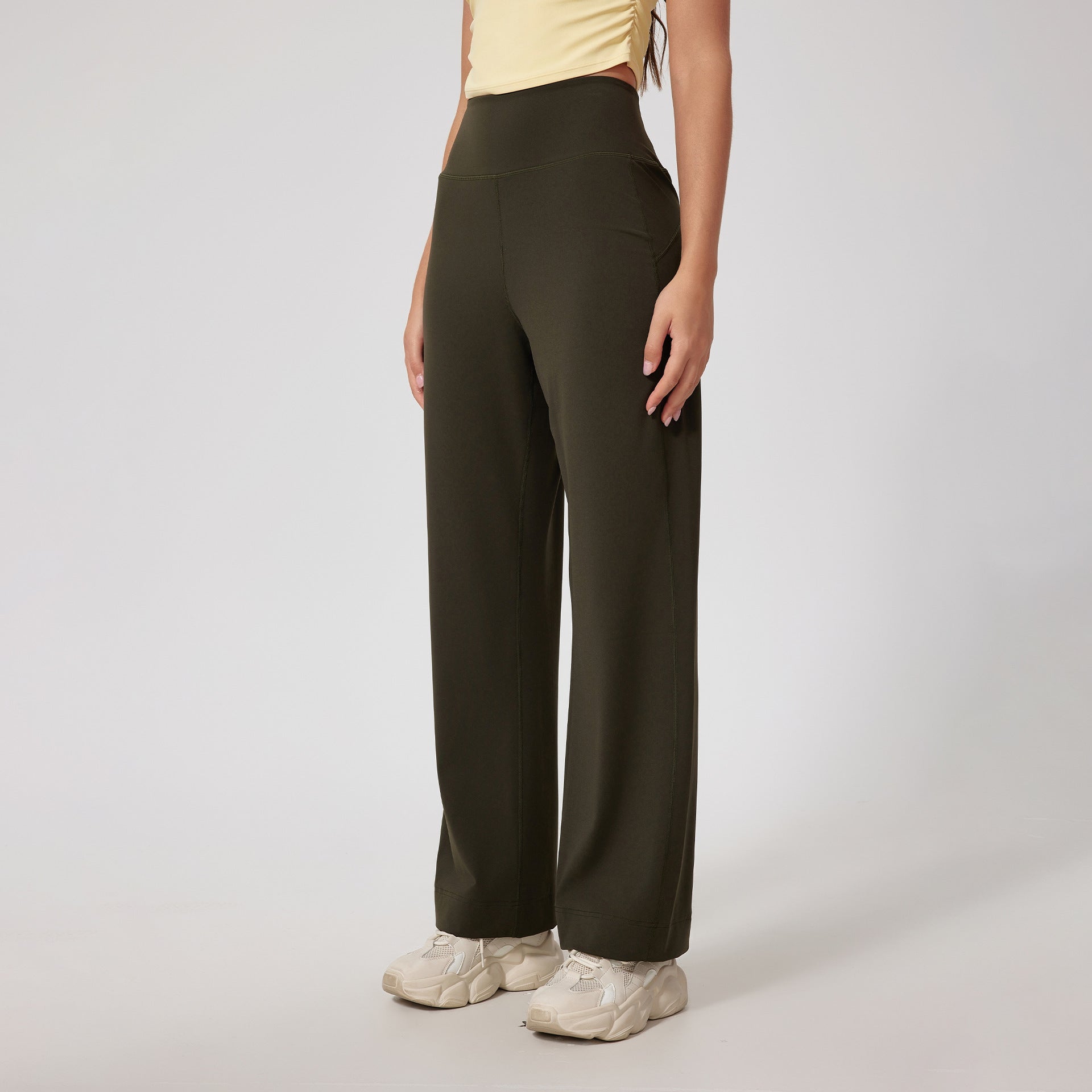 A woman wearing dark olive high-waisted wide-leg pants with a yellow crop top and cream chunky sneakers, standing against a white background