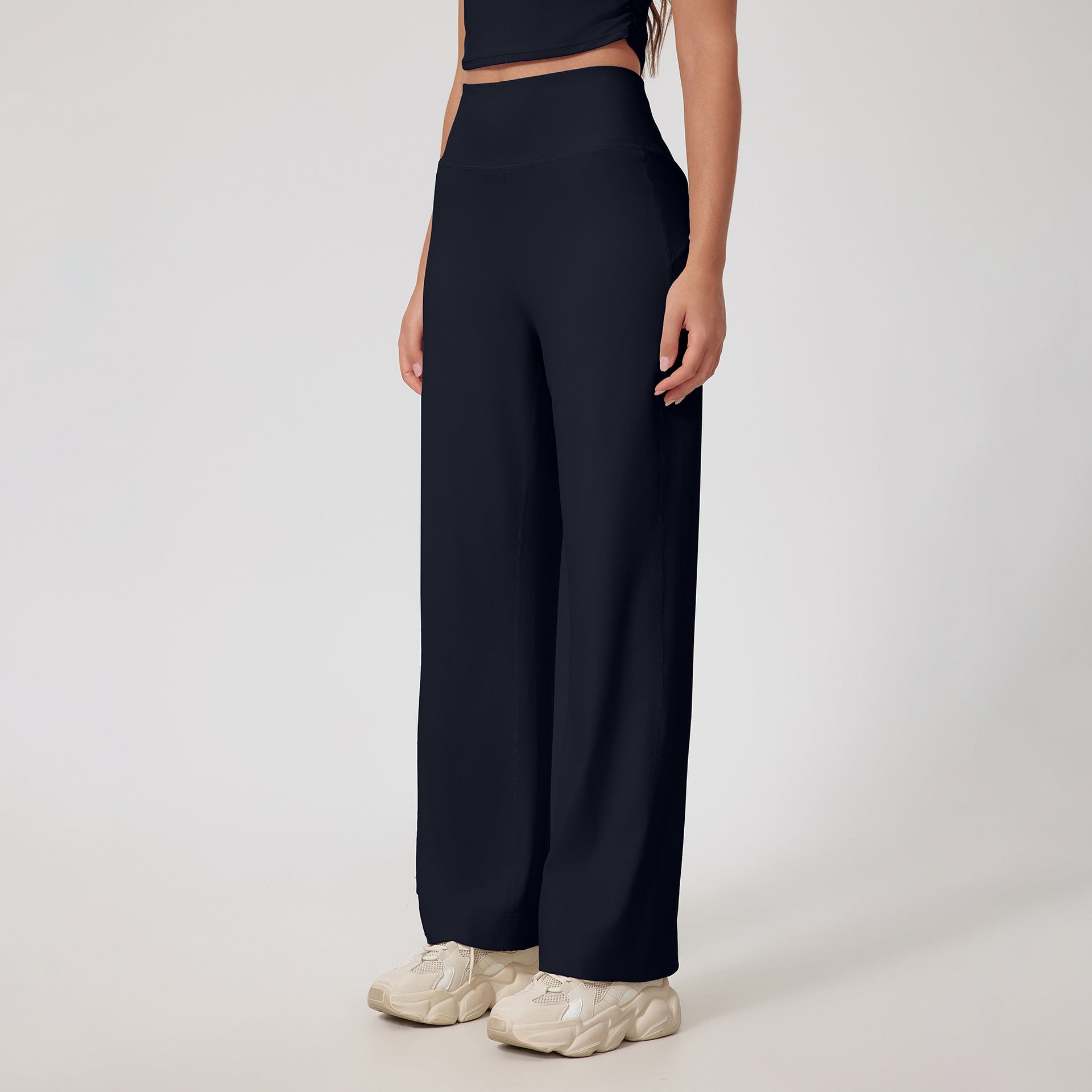A woman wearing black high-waisted wide-leg pants with a matching crop top and cream chunky sneakers, standing against a white background