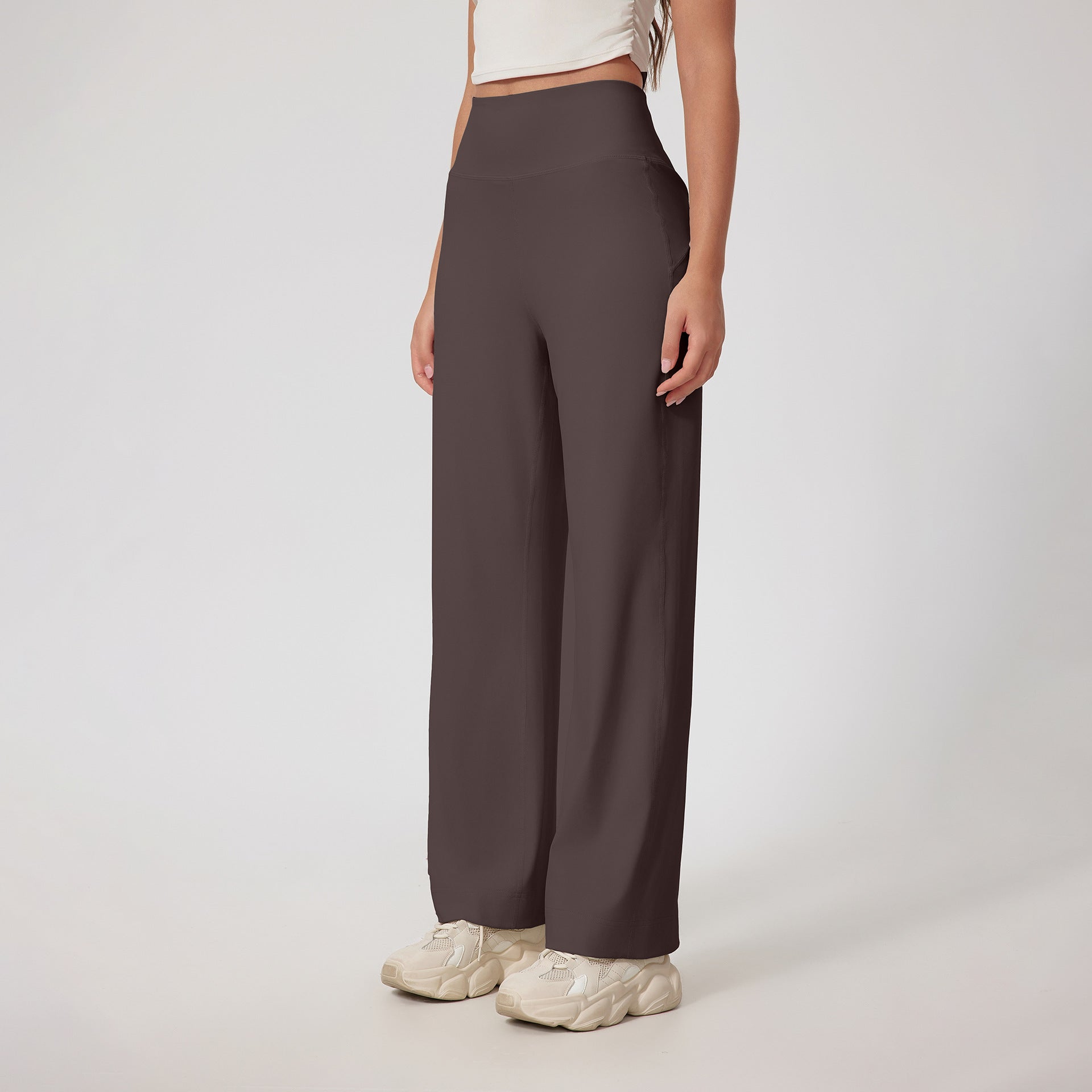 A woman wearing dark brown high-waisted wide-leg pants with a white crop top and cream chunky sneakers, posing against a white background
