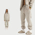 Model in cream-colored hoodie and sweatpants set with unzipped hoodie, gold watch, and beige slides against white background