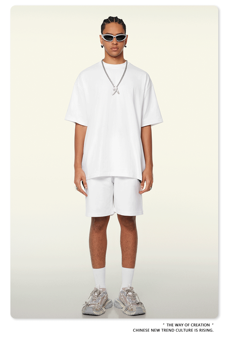 A man with braided hair wearing all-white oversized t-shirt and shorts set, styled with silver chain necklace, white sunglasses, and chunky sneakers