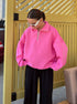 Woman in hot pink quarter-zip sweatshirt, black pants, and white sunglasses standing by yellow wall