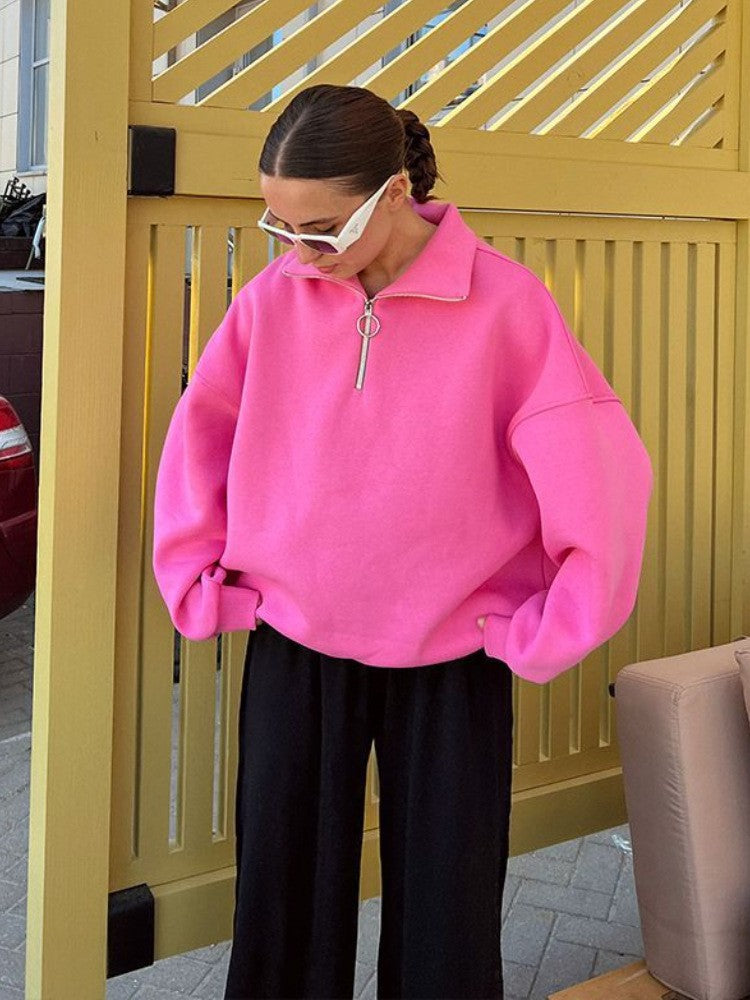 Woman in hot pink quarter-zip sweatshirt, black pants, and white sunglasses standing by yellow wall