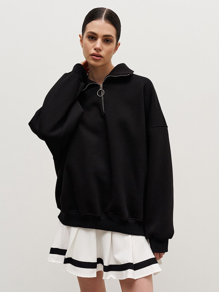 Woman wearing black oversized quarter-zip sweatshirt with ring pull and white pleated tennis skirt with black stripes