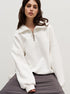 Woman in white oversized quarter-zip sweatshirt with ring pull zipper, seated on grey cushion