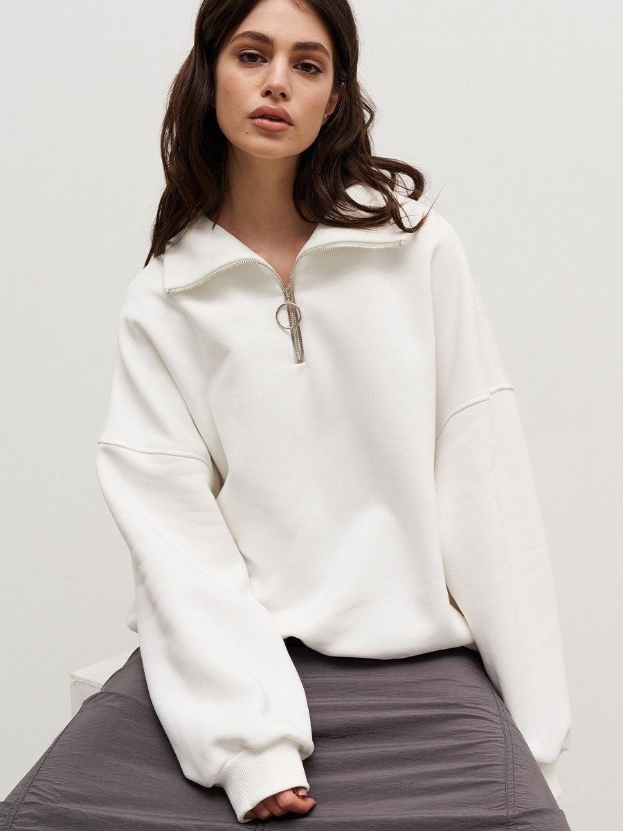Woman in white oversized quarter-zip sweatshirt with ring pull zipper, seated on grey cushion