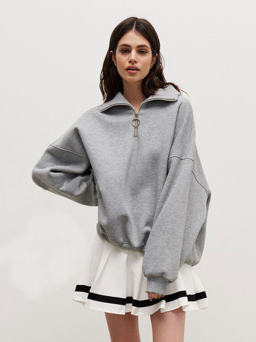 Woman in grey oversized quarter-zip sweatshirt paired with white striped-hem tennis skirt against white backdrop
