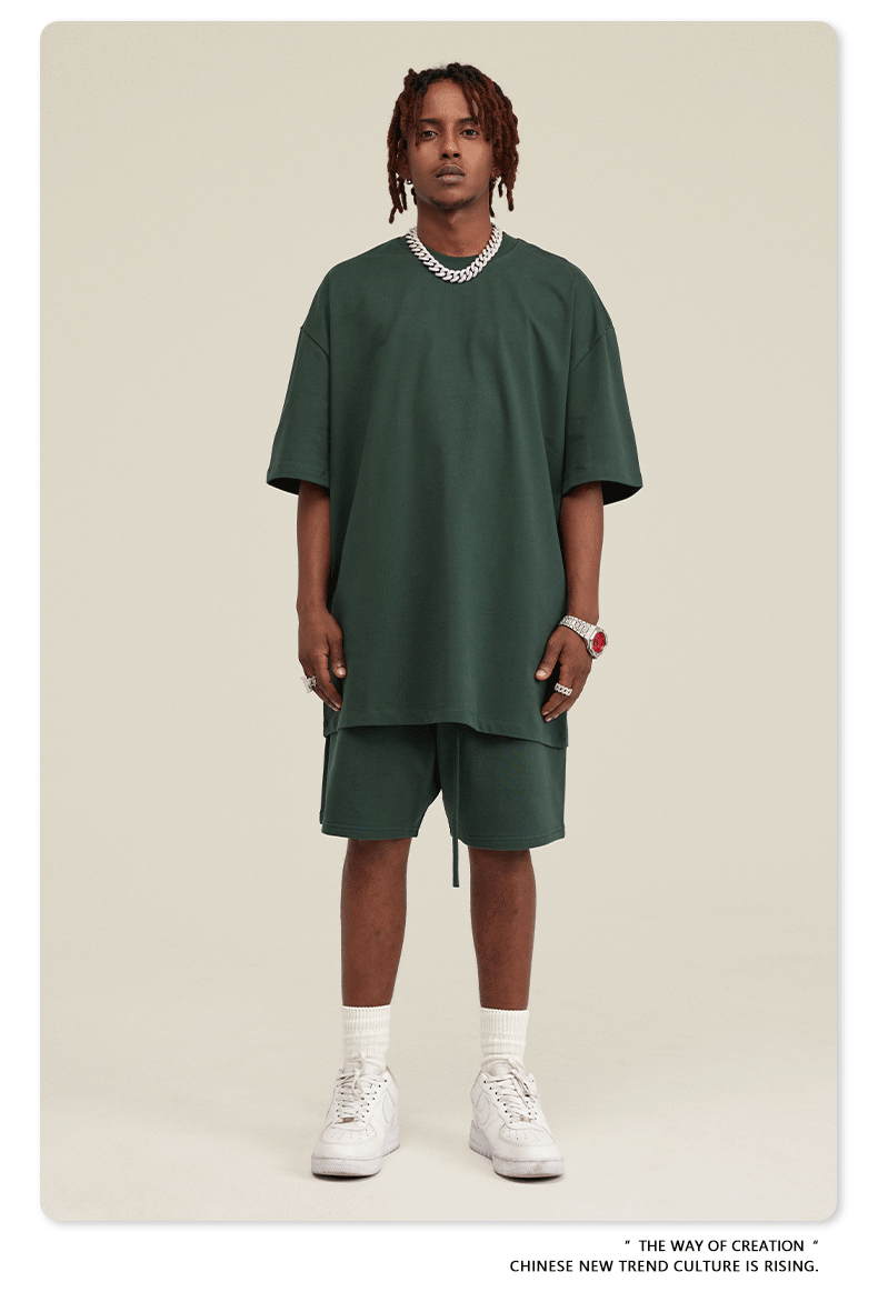 A man with short locs wearing a light sage green oversized t-shirt and matching shorts set, styled with white socks, air force ones, and a silver chain