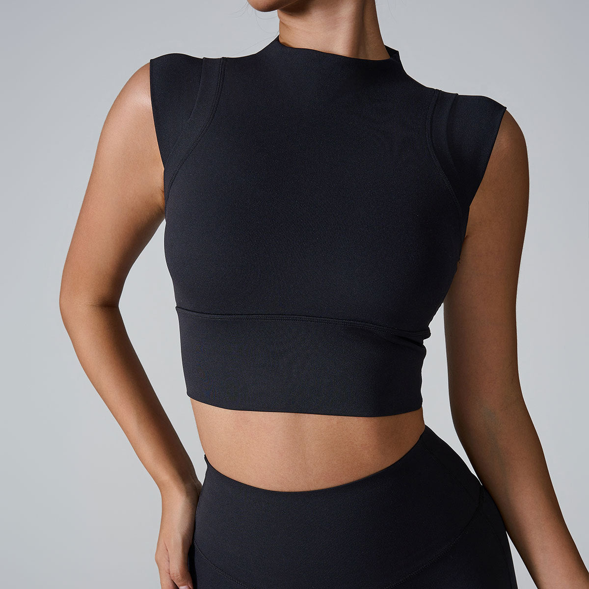 A woman is wearing a black seamless mock neck crop top paired with matching high-waisted leggings against a neutral gray backdrop